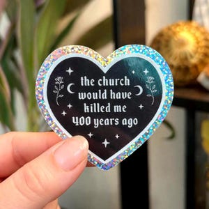 以下が含まれることがあります： ホログラフィックの縁取りと「the church would have killed me 400 years ago」の文字が入ったハート型のステッカー。ステッカーは、白い星、三日月、花のアクセントが付いた黒い背景です。