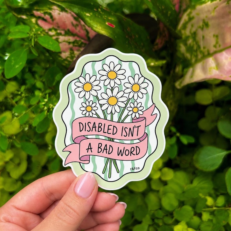 Hand holding a pastel green die-cut sticker featuring white daisies wrapped in a pink ribbon that reads “Disabled Isn’t a Bad Word” against a lush green plant background.