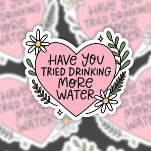 A vinyl sticker featuring a pink heart with the phrase “Have you tried drinking more water” in hand-lettered text. The heart is surrounded by white daisies, green leaves, and tiny sparkle doodles. The background is a blurred pattern of the same sticker.