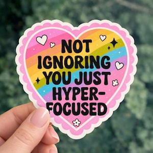 Op de afbeelding: Een hartvormige sticker met een roze geschulpte rand en een regenboogkleurig interieur. De sticker bevat de tekst "NOT IGNORING YOU JUST HYPER-FOCUSED" in zwart, met kleine decoratieve hartjes, sterren en bloemen.