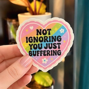 May include: A heart-shaped sticker with a pink border and a rainbow-colored interior. The sticker has the words "NOT IGNORING YOU JUST BUFFERING" in black, bold letters. Small flowers and stars decorate the design.