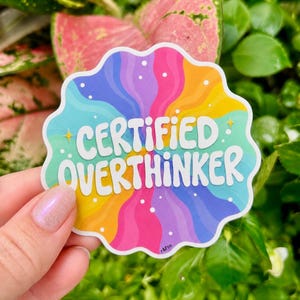 Puede incluir: Una pegatina de borde festoneado con un vibrante patrón de remolino radial en colores arcoíris. El texto "CERTIFIED OVERTHINKER" está impreso en letras blancas en negrita en el centro, acentuado por pequeños puntos blancos y estrellas.
