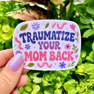 May include: A rectangular sticker with rounded corners featuring the text "TRAUMATIZE YOUR MOM BACK" in a bold, wavy, purple and pink font. The design includes small pink worms, purple and pink flowers, green leaves, and tiny sparkles.