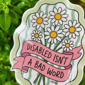 Close-up of the glossy “Disabled Isn’t a Bad Word” daisy sticker showing the handwritten typography, floral details, and shine of the waterproof vinyl finish.