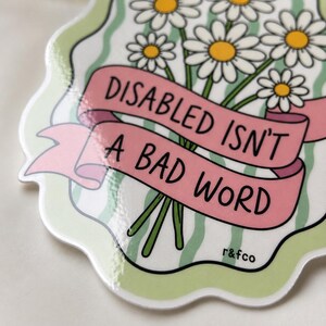 Close-up of the glossy “Disabled Isn’t a Bad Word” daisy sticker showing the handwritten typography, floral details, and shine of the waterproof vinyl finish.