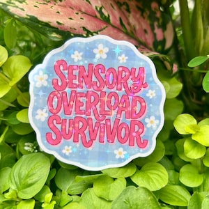Glossy holographic vinyl sticker reading “Sensory Overload Survivor” displayed among vibrant green leaves in natural sunlight. Features bold pink retro typography, a pastel blue background, white daisies, and shimmering star accents, highlighting the sticker’s neurodiversity theme, sensory overload awareness, and premium holographic vinyl finish.