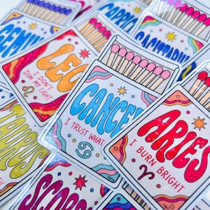 May include: A collection of rectangular stickers featuring matchbox designs with zodiac signs. Each sticker displays a sign name, a corresponding symbol, and a short phrase like I Burn Bright or I Trust What, accented with colorful wavy lines and stars.