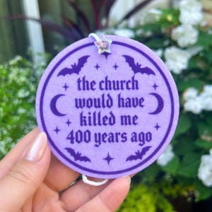 May include: A circular lavender felt ornament featuring dark purple text that reads, "the church would have killed me 400 years ago." The design includes dark purple bats, crescent moons, and stars, with a small iridescent star bead at the top.