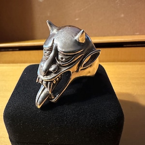 May include: A silver-toned ring featuring a detailed sculptural head of a horned creature with pointed ears, a prominent nose, and an open mouth revealing sharp teeth and a long, protruding tongue. The metal surface has a polished finish.