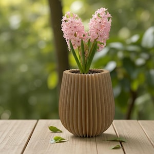 Può includere: Un vaso di fiori in legno marrone e scanalato contiene due fiori di giacinto rosa con steli verdi. Il vaso è su una superficie di legno con foglie sparse, su uno sfondo verde sfocato.