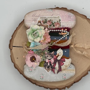 May include: A decorative spool-shaped craft piece featuring a vintage-style label with the text Madame, adorned with layered fabric flowers, sequins, a string of pearls, a kitten illustration with a pink bow, and a white swan with a red bow.