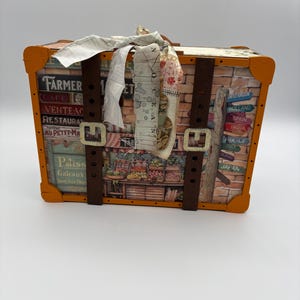 May include: A decorative wooden trunk with orange trim, featuring travel-themed imagery and text. The trunk has brown straps with silver buckles and is adorned with fabric ribbons. The text includes "Farmer Cafe" and country names.