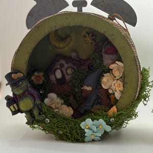 May include: A whimsical, decorative clock featuring a miniature fairy garden scene. The clock face is a circular frame with a green exterior, housing a diorama with a house, gnomes, flowers, and a frog holding a lantern. The clock has a metal top and a mossy base.