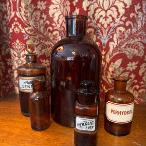 Könnte beinhalten: Fünf bernsteinfarbene Glasflaschen mit verschiedenen Etiketten. Die größte Flasche ist klar und hat eine breite Öffnung. Die anderen Flaschen sind kleiner und haben unterschiedliche Formen. Die Etiketten auf den Flaschen beinhalten die Wörter "Senna", "Hier. Glic. Fosf." und "Perhydrol".