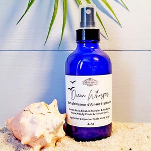 Può includere: Un flacone spray in vetro blu da 4 oz con erogatore nero, etichettato Ocean Whisper Air Freshener. L'etichetta riporta il testo Rustik Shack Shop, Aqua, Woody, Floral & Herbal Notes e Shake Well & Spray. Una conchiglia è posizionata accanto al flacone.