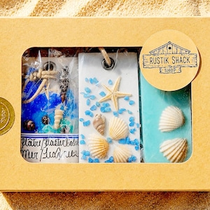 May include: A rectangular cardboard gift box containing three rectangular hanging ornaments decorated with seashells and blue accents. The box features a circular sticker with the text RUSTIK SHACK SHOP and a gold sticker reading THANK YOU.