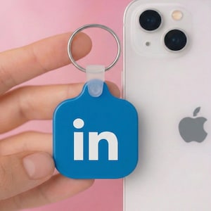 May include: A blue keychain with the LinkedIn logo, featuring the letters "in" in white, attached to a silver keyring. The keychain is held near a white smartphone with a triple-lens camera.