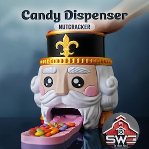 Puede incluir: Un dispensador de caramelos novedoso con forma de soldado cascanueces. El dispensador es negro, dorado y beige con barba blanca y mejillas rosadas. Los caramelos se dispensan por la boca del cascanueces. El texto "Candy Dispenser Nutcracker" está arriba.