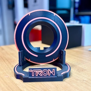 May include: A black and pink 3D-printed Tron-themed disc and stand. The disc has a circular design with a pink neon-like outline. The stand features the word "TRON" in a matching pink color. The design is reminiscent of the classic sci-fi film.