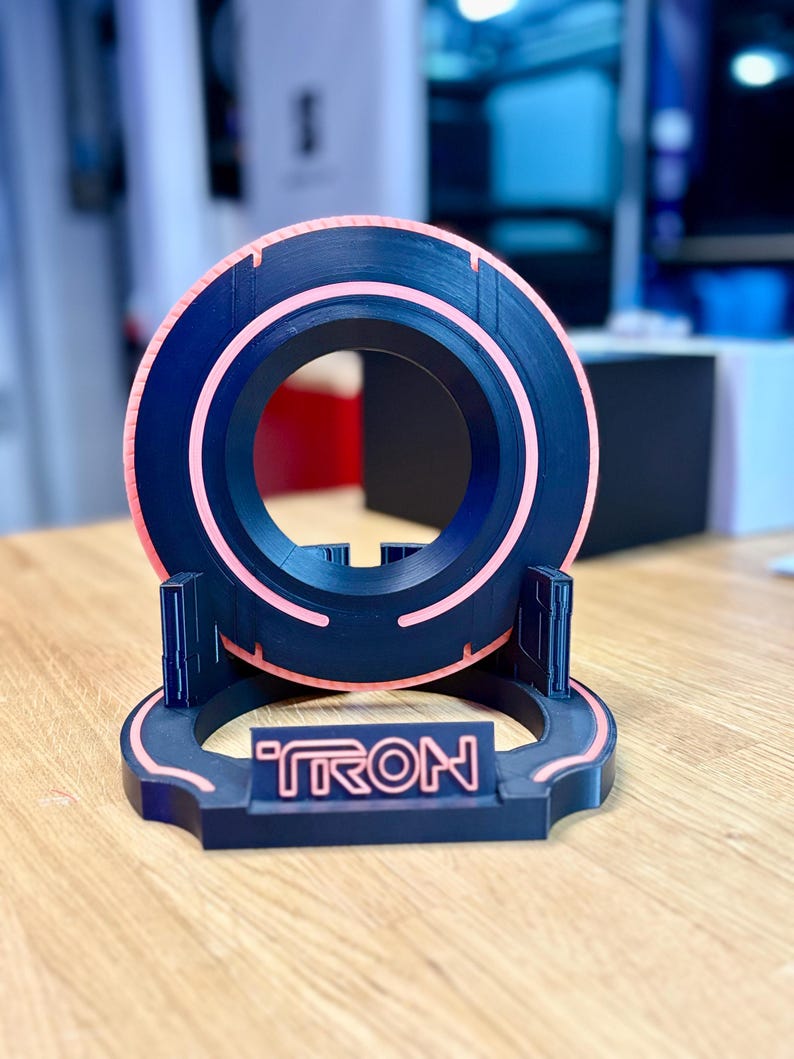 May include: A black and pink 3D-printed Tron-themed disc and stand. The disc has a circular design with a pink neon-like outline. The stand features the word "TRON" in a matching pink color. The design is reminiscent of the classic sci-fi film.