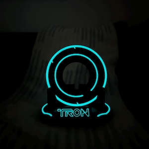 May include: A black, illuminated Tron-themed decorative object. The object features a glowing blue circular design with the word "TRON" in matching blue lettering. The design is set against a dark background.