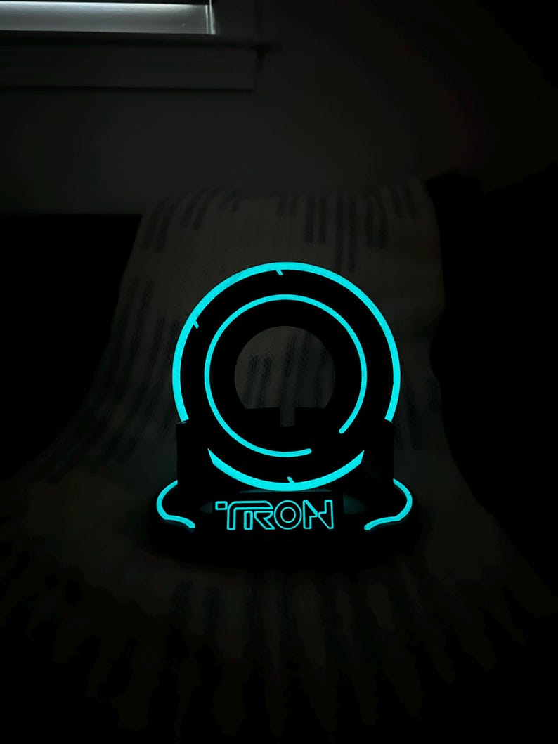 May include: A black, illuminated Tron-themed decorative object. The object features a glowing blue circular design with the word "TRON" in matching blue lettering. The design is set against a dark background.
