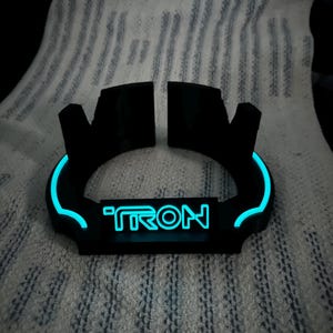 May include: Black, curved, illuminated device with the word "TRON" in blue neon. The device has a glowing blue outline and is set against a textured, white background.