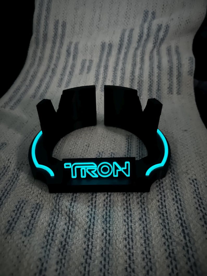 May include: Black, curved, illuminated device with the word "TRON" in blue neon. The device has a glowing blue outline and is set against a textured, white background.