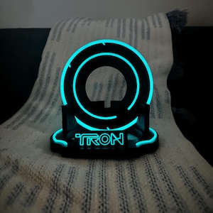 May include: Black and blue illuminated Tron-themed desk accessory. The circular design features a glowing blue outline and the word "TRON" at the base. The accessory is made of black material and sits on a white and gray patterned surface.