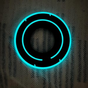 May include: A black circular object with a glowing blue outline and inner design. The object has a futuristic aesthetic, with a bright, neon-like glow. The design features a series of concentric circles, creating a sense of depth and movement. The object is set against a textured background.