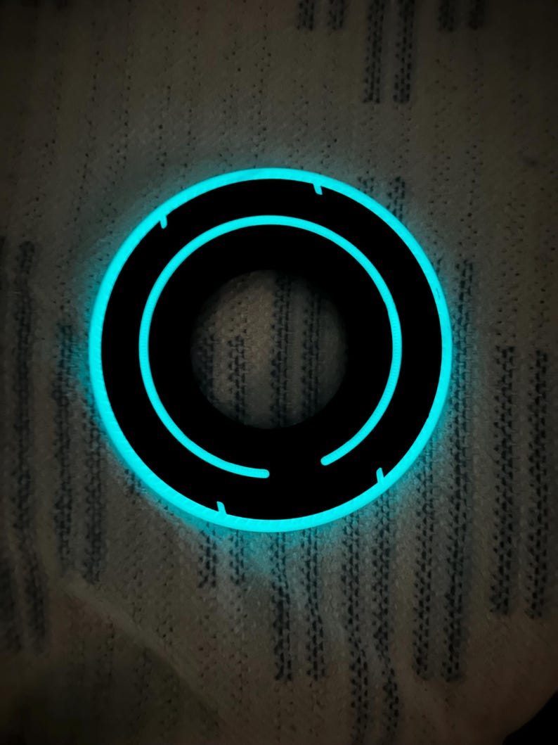 May include: A black circular object with a glowing blue outline and inner design. The object has a futuristic aesthetic, with a bright, neon-like glow. The design features a series of concentric circles, creating a sense of depth and movement. The object is set against a textured background.