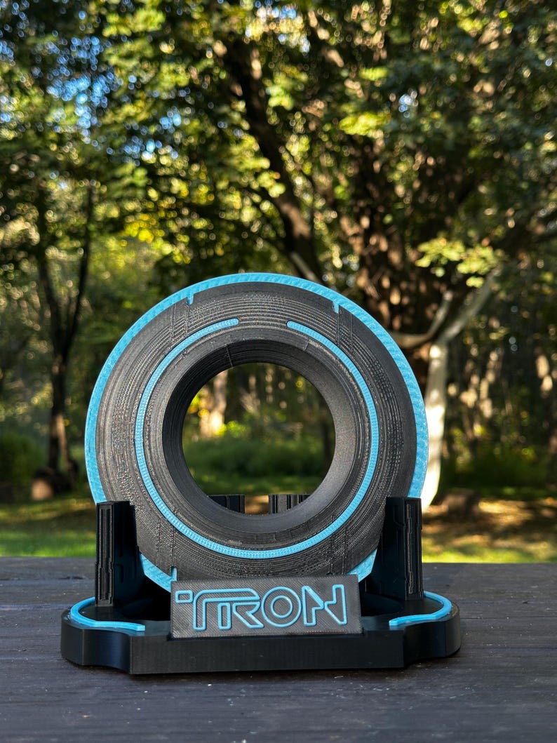 May include: A black and blue decorative disc with the word "TRON" on the base. The disc has a circular design with a blue outline and is displayed on a black stand. The background is a blurred view of trees.