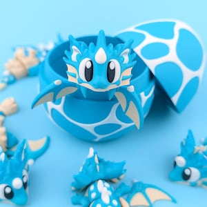 May include: A blue dragon figurine emerges from a blue and white spotted egg. The dragon has large black eyes, white accents, and tan wings. Several other dragon figurines are scattered around the egg on a light blue surface.