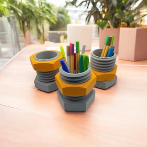 Peut inclure: Trois organiseurs de bureau gris et jaunes en forme d'écrous, chacun rempli de stylos et de crayons colorés.