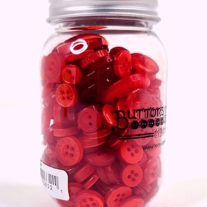 May include: A clear glass jar filled with various red buttons. The jar has a silver metal lid and a label with text and a barcode. The buttons are different shapes and sizes, with some having holes for sewing.