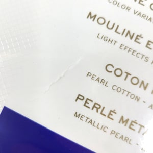 May include: Close-up of a white label with gold text that reads "Mouline'  Color Variations", "Mouline'  Light Effects", "Coton Pearl Cotton", and "Perle'  Metallic Pearl".