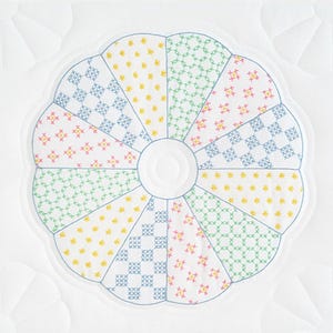 May include: A white fabric circle with a flower design in the center. The flower is made up of 12 sections, each with a different cross-stitch pattern in shades of blue, green, yellow, and pink.
