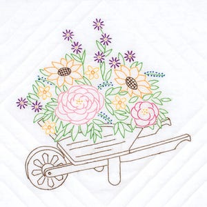 May include: A white fabric square featuring a machine embroidery design of a brown wheelbarrow filled with a colorful floral arrangement. The flowers are stitched in shades of pink, yellow, and purple with green leaves and stems on a quilted background.