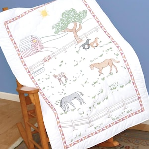 May include: White quilted fabric with a red barn, green tree, and multiple horses embroidered in different colors. The fabric has a red, white, and green border.