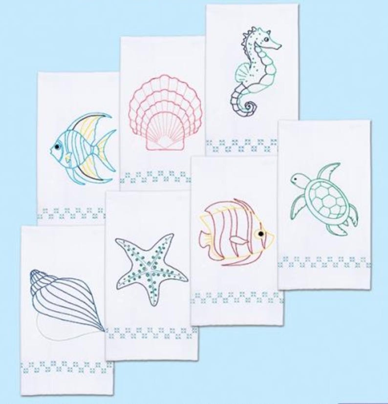 K&ouml;nnte beinhalten: Wei&szlig;e Handt&uuml;cher mit Stickereien von Meeresmotiven. Die Designs zeigen ein Seepferdchen, einen Fisch, eine Muschel, einen Seestern und eine Schildkr&ouml;te. Jedes Handtuch hat eine dekorative Bord&uuml;re. Die Stickerei verwendet blaue, gr&uuml;ne, gelbe und rote F&auml;den.