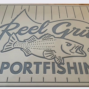 May include: A beige sign with the words "Reel Grit Sportfishing" in gray. A fish and fishing rod are also depicted. The sign has a ruler along the bottom edge. The design is carved into the sign.