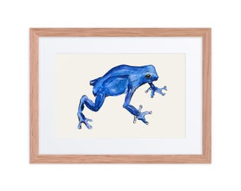 Puede incluir: Una lámina de arte enmarcada que presenta una ilustración de una rana dardo azul vibrante centrada sobre un fondo color crema. La obra está en un marco de madera clara con un paspartú blanco, mostrando a la rana de perfil lateral.