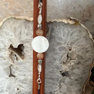 May include: A necklace featuring a central white circular shell disc flanked by two round peach-colored beads, two silver-toned metal spacers, and two elongated white shell beads, all strung on a black cord with silver-toned metal findings.