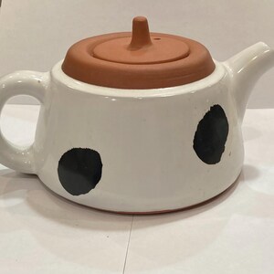 May include: White ceramic teapot with a terracotta lid and black polka dots. The teapot has a handle and a spout.