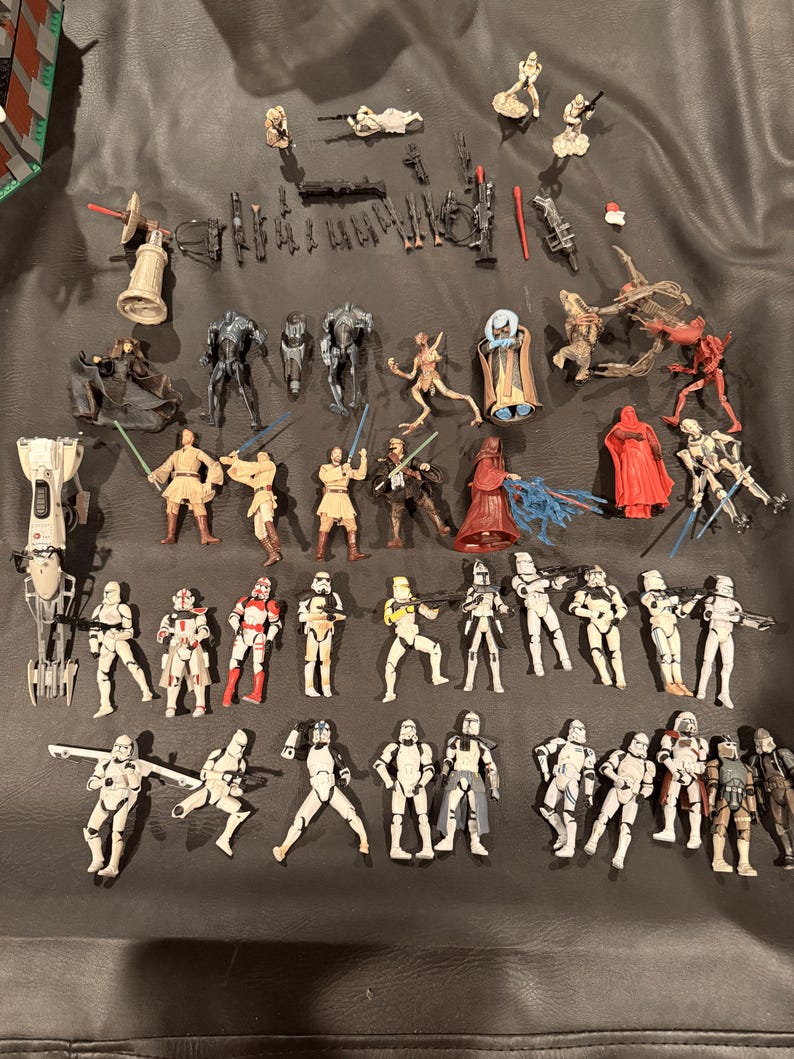 K&ouml;nnte beinhalten: Eine Sammlung von Star Wars-Actionfiguren, darunter Sturmtruppen, Jedi und verschiedene andere Charaktere. Die Figuren sind in verschiedenen Posen und weisen detaillierte Designs auf. Die Farbpalette umfasst Wei&szlig;, Schwarz, Rot und Beige.