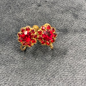 May include: A pair of vintage clip-on earrings. Each earring features a gold-toned setting with a central, round, faceted red stone surrounded by smaller red stones. The earrings have a floral design.