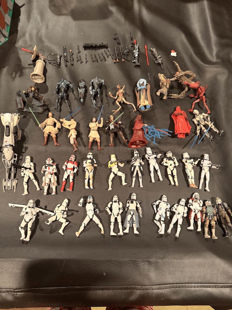 K&ouml;nnte beinhalten: Eine Sammlung von Star Wars-Actionfiguren und Zubeh&ouml;r, darunter verschiedene Charaktere und Waffen. Die Figuren sind in verschiedenen Posen und Farben, mit Fokus auf den ikonischen Charakteren der Franchise.