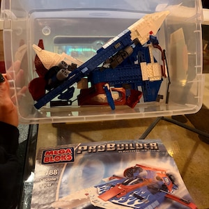 May include: A clear plastic storage bin filled with a partially constructed blue, white, and red Mega Bloks Cyclone Racer model. The instruction booklet is visible, featuring the completed model. The set is from the ProBuilder Racing Series.