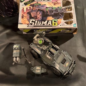 May include: G.I. Joe Sigma 6 Night Ops V.A.M.P. vehicle toy set. The box shows the vehicle in action. The toy vehicle is gray and green with black wheels. Includes additional toy components.