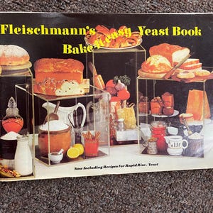 May include: A vintage cookbook cover titled "Fleischmann's Easy Yeast Book." The cover features various baked goods, including bread and pastries, displayed on clear shelves. Ingredients like flour, lemons, and jars are also visible. The text "Now Including Recipes for Rapid Rise Yeast" is at the bottom.
